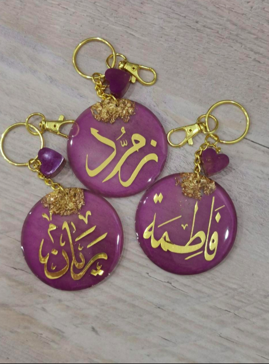 Purple keychains with gold Arabic text on a wooden surface