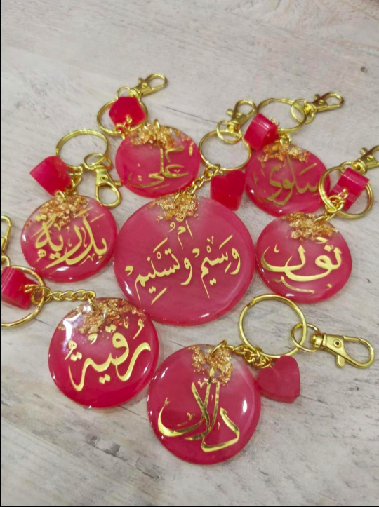 Pink keychains with gold Arabic text on a light wooden surface