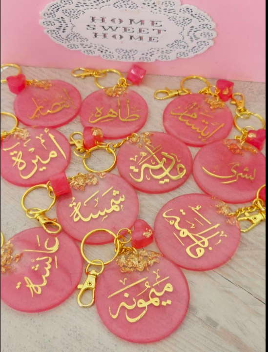 Pink keychains with gold Arabic text on a pink background with 'Home Sweet Home' sign.
