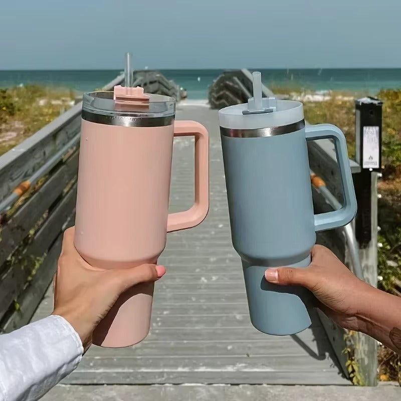 Two hands holding pink and blue insulated travel mugs on a wooden boardwalk by the sea.