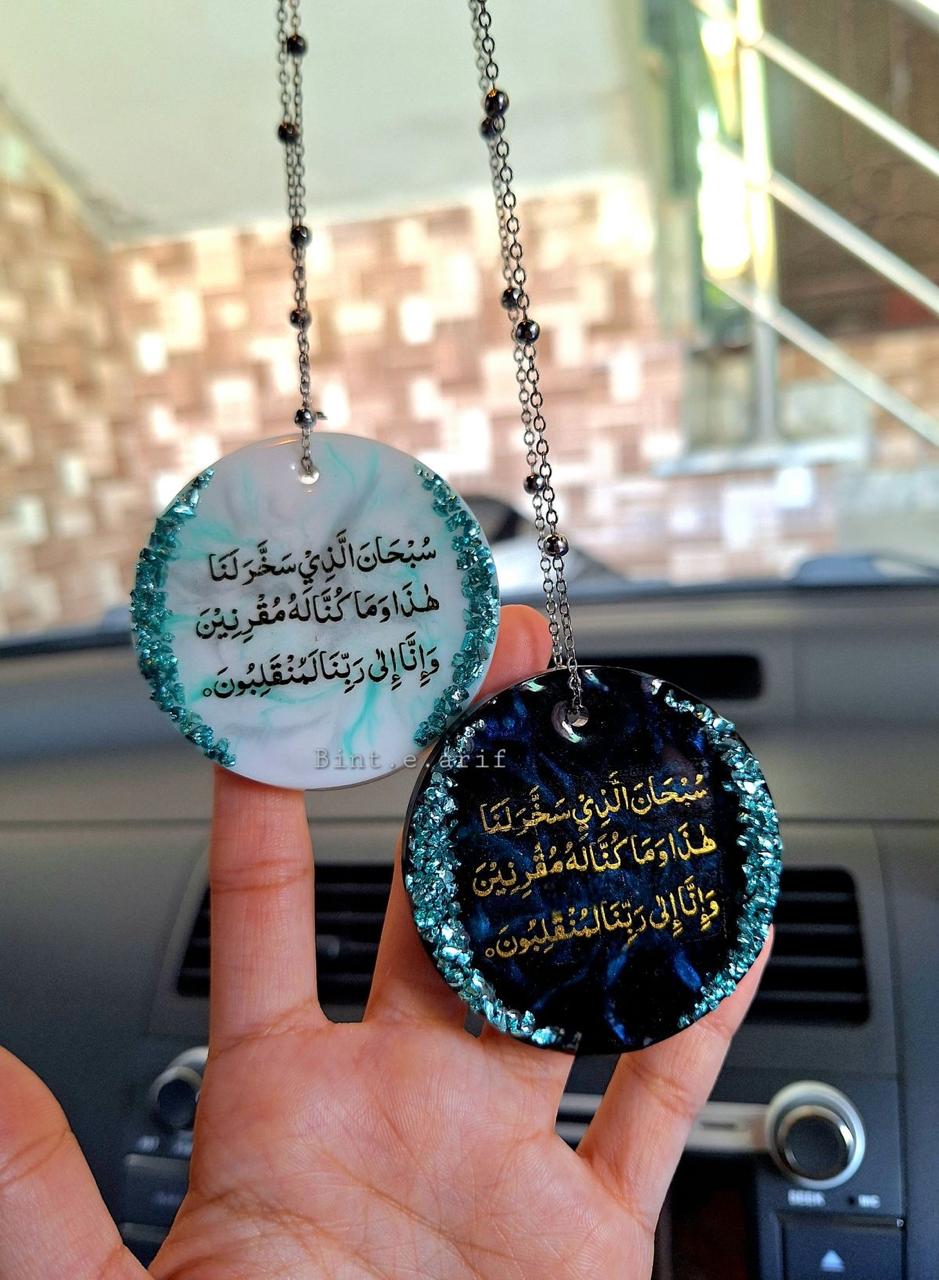 Two round hanging car decorations with Arabic text held by a hand in front of a car's interior.