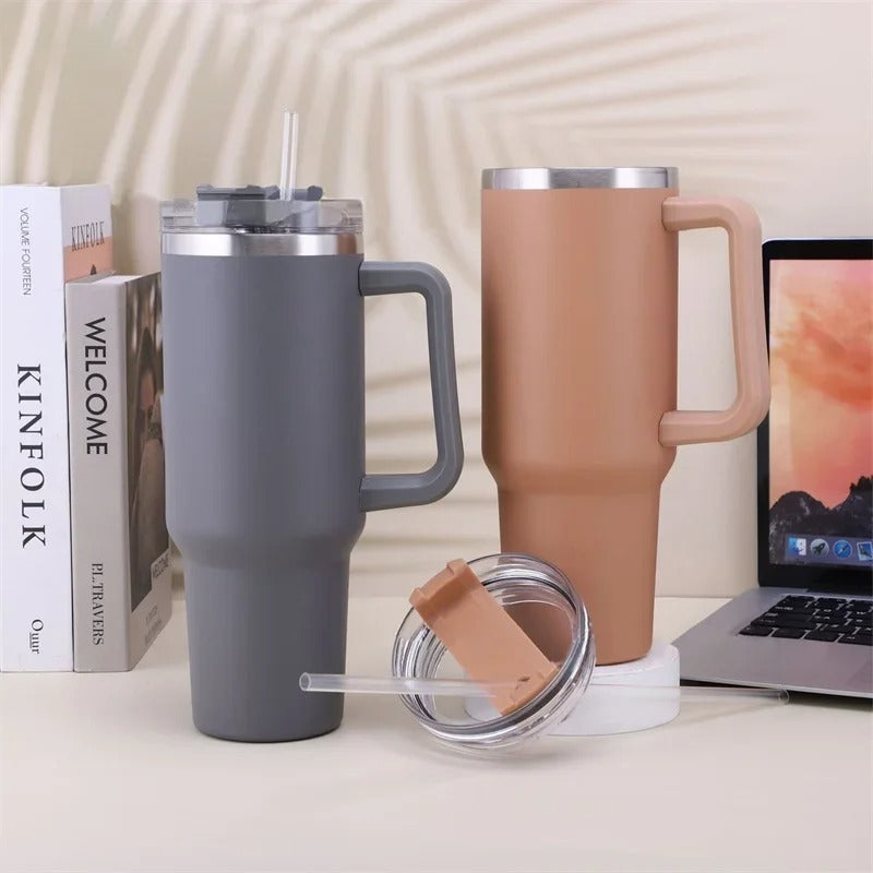 Two insulated travel mugs with handles on a surface next to a laptop and book.