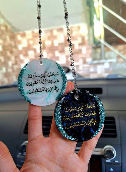 Two round hanging car decorations with Arabic text held by a hand in front of a car's interior.